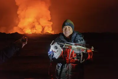 A smiling person holds a drone with a GoPro camera against a fiery volcanic eruption at night, illuminated by the glow of lava and smoke.