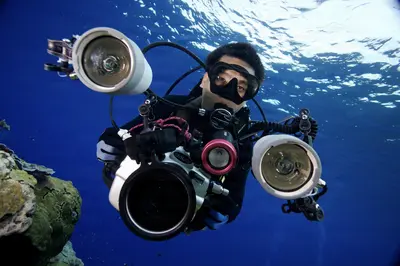 A scuba diver with a professional underwater camera rig and dual strobes captures a selfie in deep blue ocean waters, surrounded by coral and marine life.