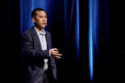 A man in a dark suit speaks on stage at a professional event, illuminated by dramatic blue lighting against a dark curtain backdrop.