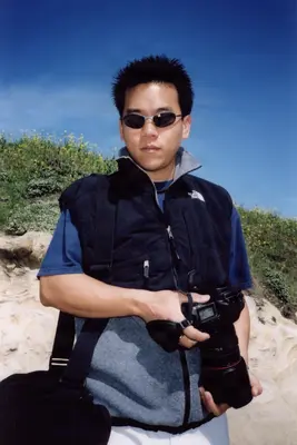 A young man in sunglasses and a vest holds a DSLR camera outdoors under a clear blue sky, posing confidently on a sunlit hillside.