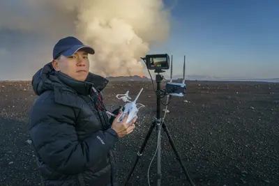 A man operates a drone controller near a tripod-mounted monitor, with a dramatic volcanic eruption billowing smoke in the background at dusk.