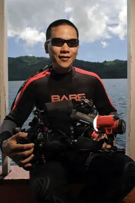 A diver in a BARE wetsuit poses with a complex underwater camera rig, including strobes and a fisheye lens, on a boat deck overlooking a tropical bay.