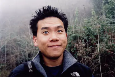 A young man with spiky hair makes a playful, wide-eyed expression while hiking in a foggy, wooded area, wearing a North Face jacket and a camera strap.