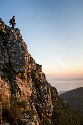 A lone hiker stands atop a rugged cliff at sunrise, silhouetted against a soft sky, overlooking a vast coastal landscape.