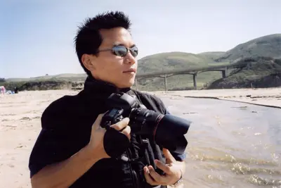 A young man in sunglasses holds a DSLR camera on a sunlit beach, with a bridge and green hills in the background.