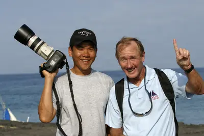 Two smiling men pose by the ocean at dawn, one holding a DSLR with a large lens, the other pointing upward, capturing a joyful moment of adventure and photography.