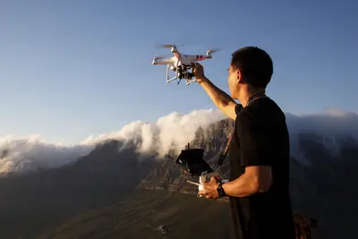 A man operates a drone at sunrise atop a mountain, with clouds swirling around rugged peaks below.
