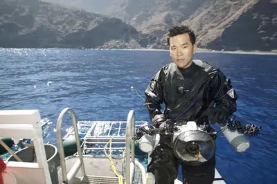 A diver in full gear stands on a boat deck at Guadalupe Island, Mexico, holding a large underwater camera, ready for a great white shark dive.