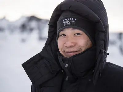 A smiling man in a black puffer jacket and beanie stands in a snowy Icelandic landscape, with snow-covered mountains in the soft light of a winter afternoon.